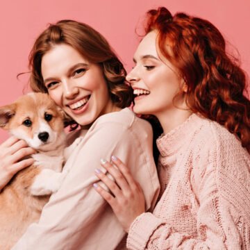 Wonderful girls holding cute dog isolated on pink background. Studio shot of smiling european ladies posing with pet Wonderful girls holding cute dog isolated on pink background. Studio shot of smiling european ladies posing with pet.